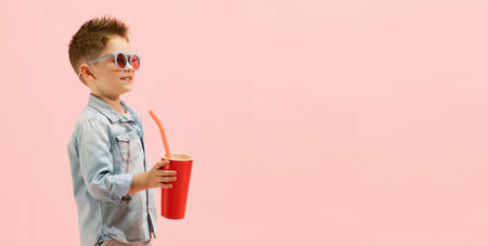 Portrait of stylish little boy, child in jeans t-shirt and sunglasses, drinking lemonade isolated over pink background. Summertime mood. Concept of childhood, family, emotions, lifestyle, fashion, adの写真素材
