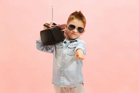 Portrait of stylish little boy, child in jeans t-shirt and sunglasses posing, listening to radio isolated over pink background. Concept of childhood, family, emotions, lifestyle, fashion, adの写真素材