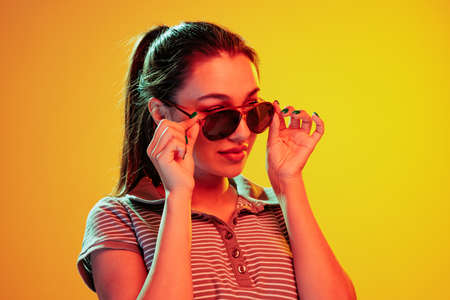 Portrait of beautiful young girl posing in stylish sunglasses isolated over yellow background in neon light. Concept of emotions, facial expression, lifestyle, fashion, youth cultureの写真素材