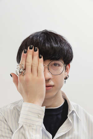 Portrait of stylish young man with piercing, in glasses, covering half face with hand, posing...