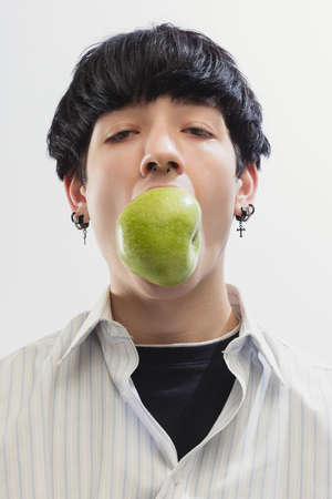 Portrait of young man in shirt posing with apple isolated over grey studio background. Stylish accessories look. Concept of modern fashion, art photography, style, queer, uniqueness, adの写真素材