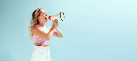 Portrait of young cheerful woman shouting in megaphone isolated over light blue studio background. Summertime sales. Concept of beauty, emotions, facial expression, lifestyle, fashion, youth cultureの写真素材