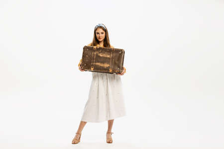 Portrait of stylish, beautiful, young woman posing with vintage suitcase isolated over white studio background. Concept of retro fashion, style, youth culture, emotions, beauty, adの写真素材