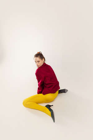 Portrait of young stylish girl in yellow tights, red jacket and heel shoes, sitting, posing isolated over grey studio background. Concept of retro fashion, art photography, style, queer, beautyの写真素材