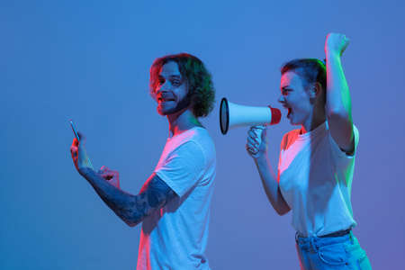 Young man working on tablet and woman shouting in megaphone isolated over blue background in neon...