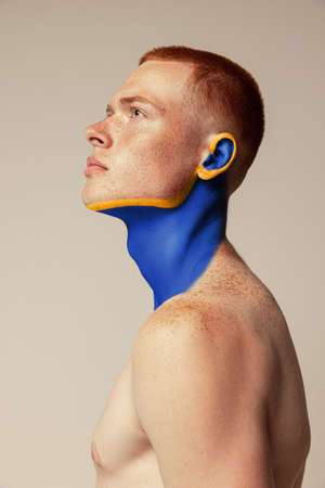 Portrait of handsome freckled man with yellow and blue drawing element on face posing, looking away isolated on grey background. Body art, aesthetics. Concept of creativity, beauty, fashion, youthの写真素材