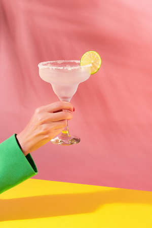 Female hand holding glass of delicious margarita cocktail isolated over pink yellow background. Cheers. Concept of cocktails, alcoholic drinks, taste, party, mix. Copy space for ad. Retro styleの写真素材