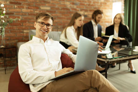 Portrait of cheerful young manager working on laptop at stylish office. Assistance of team. Young people working together on project making and launching. Concept of success and businessの写真素材