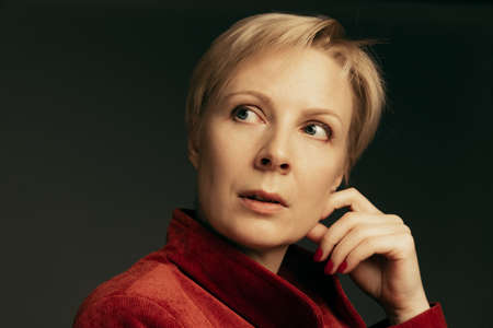 Portrait of beautiful middle-aged woman in stylish red velvet jacket posing isolated over dark background. Thoughtful, dreamy look. Concept of beauty, fashion, style, live portrait. Copy space for adの写真素材