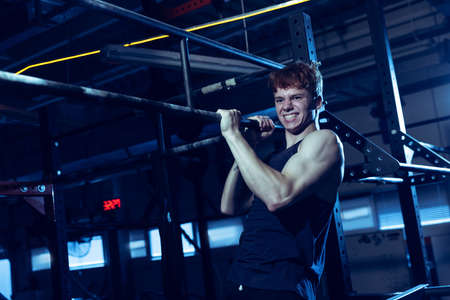 Portrait of sportive muscular young man training, doing pull up exrcises isolated over gym...