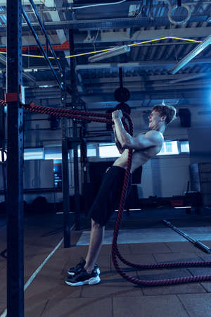 Full-length portrait of muscular red-haired man training, doing exercises with ropes isolated over...