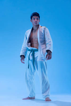 Portrait of young sportive man, judo athlete posing in white kimono with green belt isolated over blue background in neon light. Concept of martial art, combat sport, health, strength, energy, fit.の写真素材
