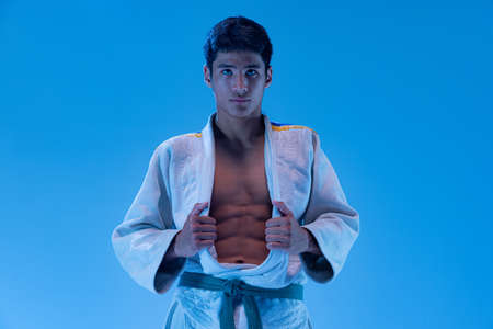 Portrait of young sportive man, combat sport athlete posing isolated over blue studio background in neon light. Concept of martial art, combat sport, health, strength, energy, fit. Copy space for adの写真素材