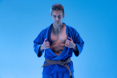 Portrait of young sportive man in blue kimono posing isolated over blue background in neon light. Concentrated. Concept of martial art, combat sport, health, strength, energy, fit. Copy space for adの写真素材