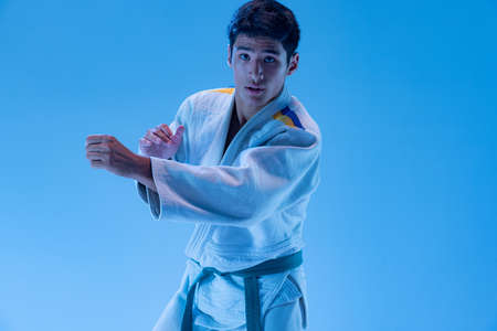 Portrait of young sportive man in white kimono posing, ready to punch isolated over blue studio background in neon light. Concept of martial art, combat sport, health, strength, energy, fit.の写真素材