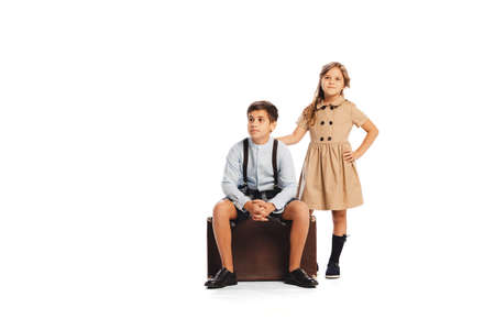 Portrait of stylish children in retro styled outfits posing, sitting on retro suitcase isolated on white studio background. Travelling. Concept of childhood, friendship, fun, lifestyle, fashion, styleの写真素材