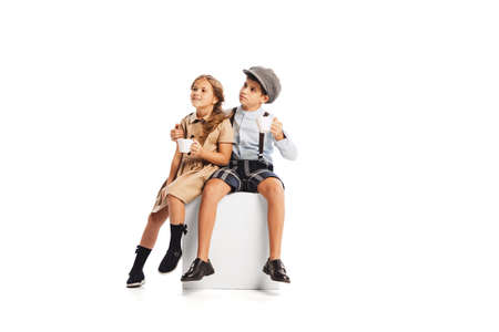 Portrait of lovely children, boy and girl in stylish retro clothes sitting together, drinking tea isolated over white background. Concept of childhood, friendship, fun, lifestyle, fashion. Retro styleの写真素材