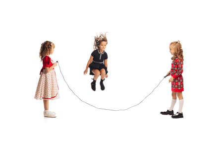 Portrait of little girls, children playing together, jumping over a rope isolated on white studio background. Concept of childhood, friendship, fun, lifestyle, fashion. Retro styleの写真素材