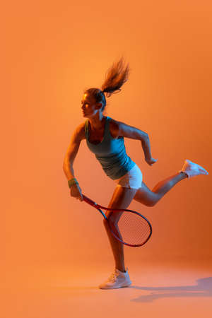 Sportive woman, professional tennis player ion action, training isolated over orange studio background in neon light. Concept of sport, health, strength, motion, hobby, lifestyle. Copy space for adの写真素材