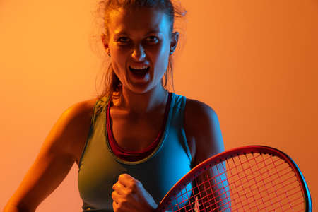 Portrait of emotive woman, professional tennis player posing isolated on orange studio background in neon light. Concept of sport, health, strength, action, motion, hobby, lifestyle. Copy space for adの写真素材