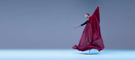 Portrait of young ballerina dancing covered with deep red transparent fabric isolated over blue grey studio background. Concept of classic ballet, inspiration, beauty, dance, creativityの写真素材