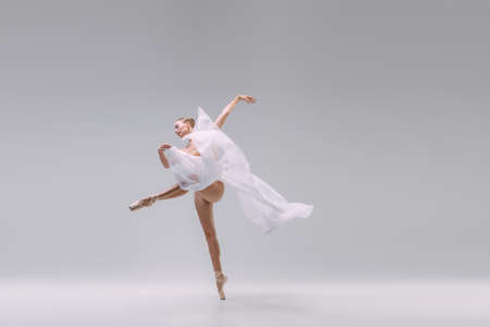 Portrait of young gracefull ballerina dancing with transparent white fabric isolated on grey studio background. Beautiful performance. Concept of classic ballet, inspiration, beauty, dance, creativityの写真素材