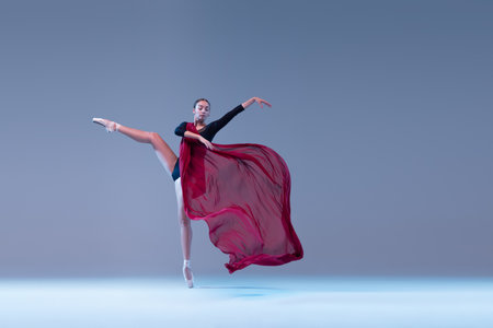 Portrait of young ballerina dancing with deep red transparent fabric isolated over blue grey studio background. Passion, femininity. Concept of classic ballet, inspiration, beauty, dance, creativityの写真素材