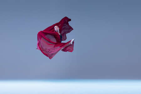 Portrait of young ballerina dancing covered with deep red transparent fabric isolated over blue grey studio background. Flying high. Concept of classic ballet, inspiration, beauty, dance, creativityの写真素材