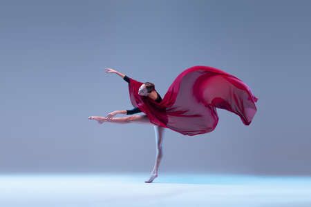 Portrait of young graceful ballerina dancing with deep red transparent fabric isolated over blue grey studio background. Tenderness. Concept of classic ballet, inspiration, beauty, dance, creativityの写真素材