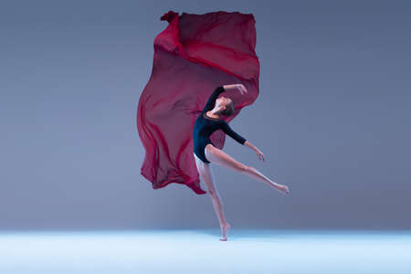 One young tender ballerina dancing with deep red transparent fabric isolated over blue grey studio background. Attractive performance. Concept of classic ballet, inspiration, beauty, dance, creativityの写真素材