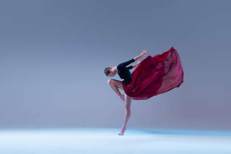 Portrait of young ballerina dancing in black bodysuit with deep red fabric isolated on blue grey...