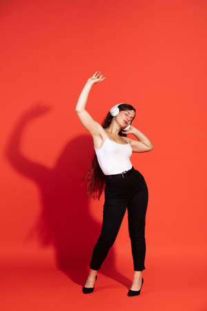 Portrait of beautiful young woman posing, listening to music in headphones, dancing isolated over...