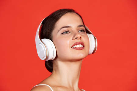 Close-up portrait of beautiful young woman posing in white headphones isolated over red studio background. Stereo system. Concept of music, education, lifestyle, youth, emotions, facial expression, adの写真素材
