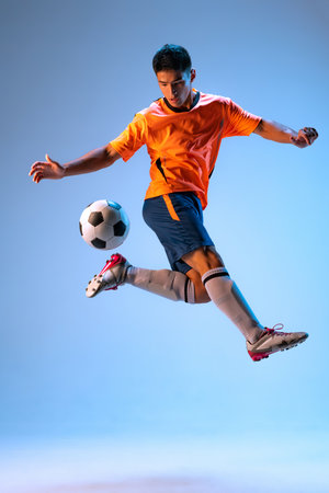 Portrait of young man, football player in motion, training, kicking ball in a jump isolated over blue background in neon light. Concept of sport, team game, action, motion. Copy space for ad, posterの写真素材