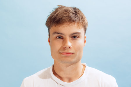 Close-up portrait of young man in white casual T-shirt looking at camera, posing isolated over blue...