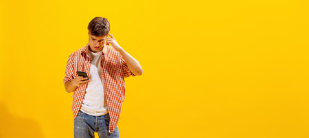 Portrait of young man in checkered shirt posing, looking in phone with curious expression isolated over yellow studio background. Concept of emotions, facial expression, lifestyle, fashion, youthの写真素材