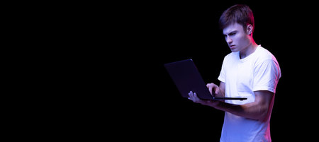 Portrait of young man in white T-shirt posing, working in laptop isolated over black background in...
