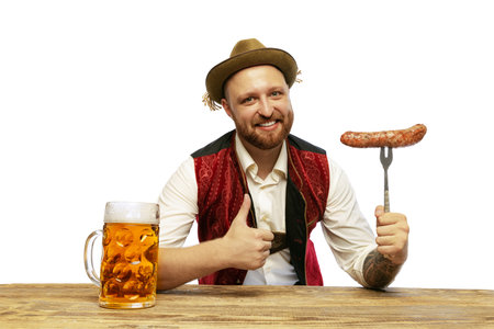 Portrait of young man wearing traditional Bavarian or German clothes with sausage and beer isolated over white background. Alcohol, Oktoberfest festival, traditions, taste concept. Copy space for adの写真素材