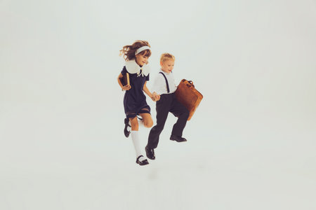 Portrait of cheerful pupils, boy and girl in school uniform running, jumping isolated over grey background. Concept of education, childhood, friendship, fun, lifestyle, fashion, retro styleの写真素材