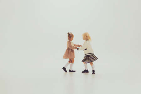 Portrait of little beautiful girls, children holding hands and spinning, playing isolated over grey studio background. Concept of childhood, friendship, fun, lifestyle, fashion, retro styleの写真素材