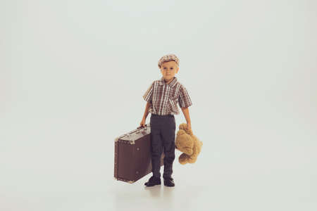 Portrait of little boy, child in checkered shirt posing with vintage suitcase and bear toy isolated over grey studio background. Concept of childhood, friendship, fun, lifestyle, fashion, retro styleの写真素材