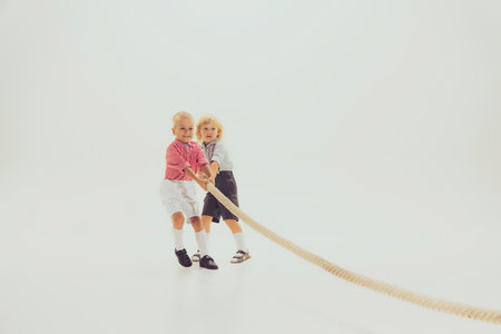 Portrait of two boys, children playing together, pulling the rope isolated over grey studio background. Game. Concept of childhood, friendship, fun, lifestyle, fashion, retro styleの写真素材