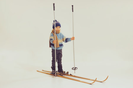 Portrait of little boy, child in winter clothes, jacket and hat standing on skis isolated over grey studio background. Winter holidays. Concept of childhood, fun, lifestyle, fashion, retro styleの写真素材