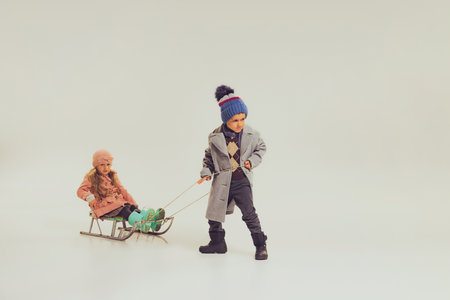 Portrait of boy and girl, children in winter clothes sledding isolated over grey studio background. Winter holidays. Concept of childhood, friendship, fun, lifestyle, fashion, retro styleの写真素材