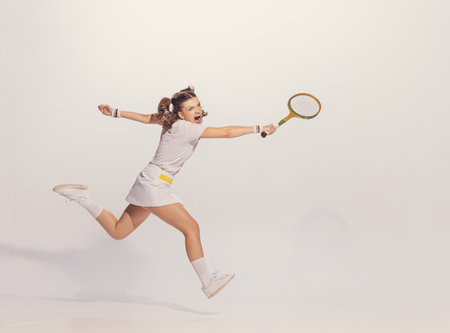 Portrait of young emotive girl in white uniform playing badminton isolated over grey studio background. Concept of emotions, sport, hobby, active lifestyle, retro fashion, beauty. Copy space for adの写真素材
