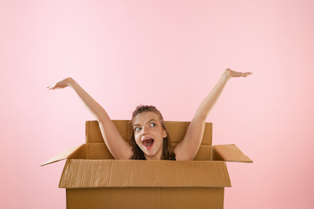 Young woman sitting inside open cardboard box with cheerful and happy expression. Moving into new apartment. Delivery service. Concept of gifts, shopping, emotions, facial expression. Unpacking box.の写真素材