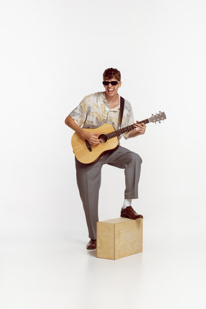 Portrait of young ma in sunglasses playing guitar, posing isolated over white studio background....