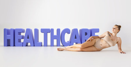 Portrait of tender young woman lying on floor on beige lingerie over 3D healthcare lettering on grey background. Natural beauty, Concept of skincare, body care, cosmetology, plastic surgery, wellnessの写真素材