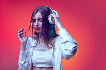 Portrait of young woman posing, listening to loud music on headphones isolated over red background in neon light. Concept of beauty, lifestyle, youth, emotions, facial expression, ad.の写真素材