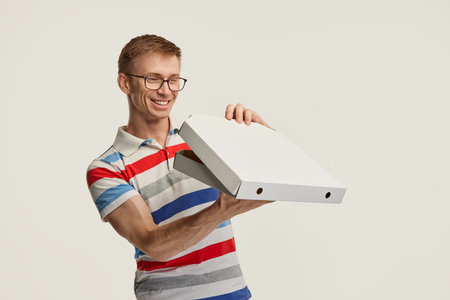 Portrait of young man in glasses getting pizza delivery, opening box isolated over white studio...